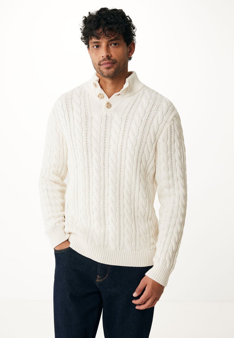 Pull en ivoire tricoté avec un motif en cable, comprenant un col haut, deux boutons et des poignets et un ourlet côtelés. Porté avec un jean foncé.