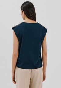 Marineblaues ärmelloses Oberteil mit rundem Ausschnitt, das mit strukturierten Schulterakzenten und einer lockeren Passform versehen ist. Trägt dazu hellbeige Hosen.