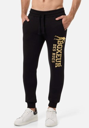 Schwarze Jogginghosen mit einem Kordelzug an der Taille, versehen mit einem goldenen Logo-Druck am linken Bein. Das Material wirkt weich und hat gerippte Bündchen.