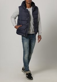 Mann trägt eine marineblaue Steppweste über einem grauen Sweatshirt, blauen Jeans und schwarzen Sneakern, steht mit einer Hand in der Tasche vor einem einfarbigen Hintergrund.