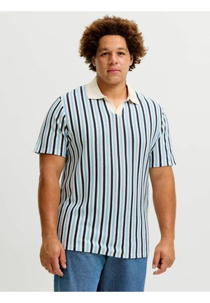 Homme aux cheveux bouclés portant une chemise à manches courtes rayée verticalement bleue et blanche et un pantalon bleu, debout devant un fond uni.