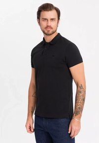 Černé polo tričko s límečkem, krátkými rukávy a decentním logem na hrudi. Materiál z bavlněné směsi s postranními rozparky pro pohodlí.