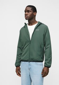 Άντρας που φοράει πράσινο μπουφάν με κουκούλα και λογότυπο EA7 πάνω από λευκό πουκάμισο και ανοιχτόχρωμο τζιν, στέκεται μπροστά σε απλό φόντο.