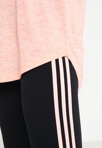 T-shirt rose clair mélangé avec fentes latérales, accompagné de leggings noirs ornés de trois bandes verticales roses le long des côtés extérieurs.