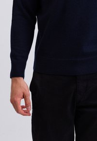 Námořnická modrá, měkká pletená svetr s dlouhými rukávy a žebrovanými manžetami, spojený s černými texturovanými kalhotami; ruka položená na boku.