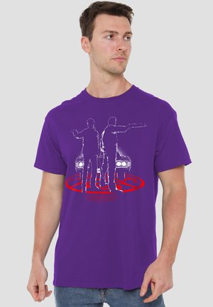 Man wearing purple t-shirt with white silhouettes of two men holding guns in front of a car and red circular symbol below.