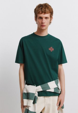 Młody mężczyzna o kręconych włosach, ubrany w ciemnozielony t-shirt i jasne spodnie, z pasiastym swetrem związanym w pasie na jednolitym tle.