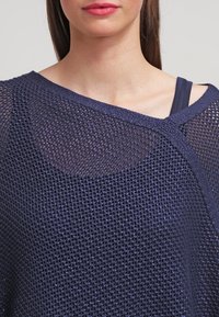 Mörkblå, löst stickad tröja med en asymmetrisk halsringning och texturerad väv. Över en figurnära linne i mörk färg.