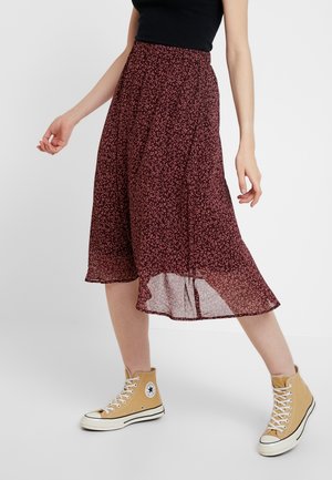 Vrouw draagt een bordeauxrode midi-rok met patroon en een split, een zwarte mouwloze top en beige hoge sneakers, staand tegen een witte achtergrond.