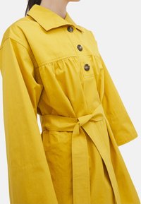 Abito di cotone giallo con scollo a colletto, bottoni in legno, maniche lunghe e cintura da annodare. Presenta una vita arricciata e tasche laterali.