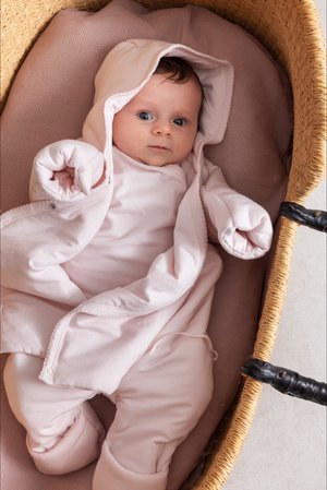 Combinaison rembourrée rose pâle avec une capuche, des manches repliées et une fermeture boutonnée, dans un panier tissé doublé d'un tissu doux et texturé.