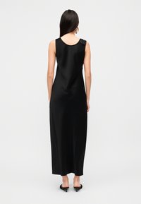 Μαύρο μακρύ φόρεμα από σατέν με στρογγυλή λαιμόκοψη, που διαθέτει μικρή βολάν λεπτομέρεια σε έναν ώμο και λεία, ρέουσα σιλουέτα.