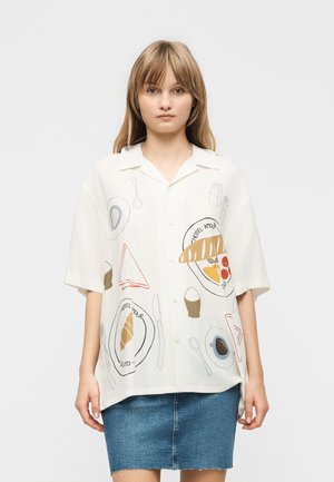 Junge Frau trägt ein weißes Kurzarmshirt mit frühstücksbezogenen Illustrationen und einen blauen Jeansrock vor einfachem Hintergrund.