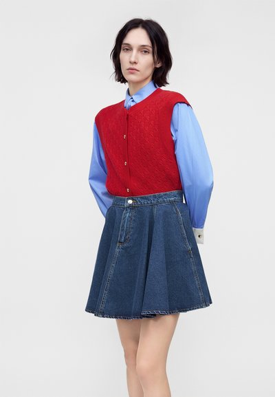 Femme aux cheveux courts et foncés porte un gilet rouge tricoté sur une chemise bleu clair à boutons et une jupe plissée en jean, debout devant un fond uni.