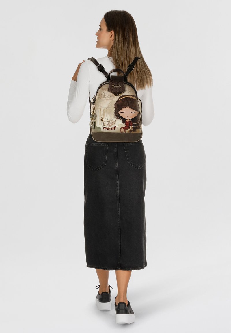 Rucksack mit einem Cartoon-Design auf einer strukturierten Oberfläche, hauptsächlich in Erdtönen, mit verstellbaren Riemen und angehängten Anhängern.