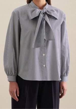 Femme portant un chemisier à manches longues, rayé noir et blanc avec un grand nœud au col, et un pantalon noir.