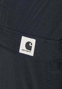 Dark navy fabric with a smooth texture features a visible white square label with a black logo on the left side, indicating the brand Carhartt.
