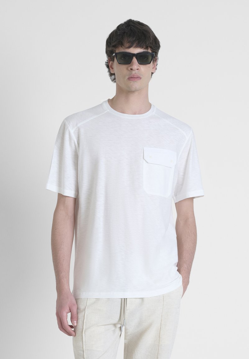 T-shirt bianco a maniche corte con scollatura rotonda, taschino frontale e tessuto testurizzato. Abbinato a pantaloni chiari.
