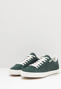 Baskets en toile verte avec lacets blancs, bout arrondi, panneaux latéraux texturés et semelle en caoutchouc. Étiquette de marque minimale sur le côté.