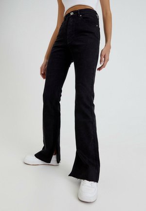 Kobieta ubrana w czarne dzwony z wysokim stanem i przednimi rozcięciami, białe trampki oraz krótki biały top na jednolitym tle.