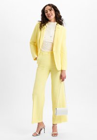 Gele op maat gemaakte pakken met een getailleerde blazer en brede pantalons, gecombineerd met een crèmekleurige coltrui. Een zilveren handtas en sandalen met straps maken de look compleet.