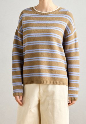 Lysebrun sweater med lyseblå vandrette striber, båret med cremefarvede bukser med brede ben på en person, der står mod en ensfarvet baggrund.