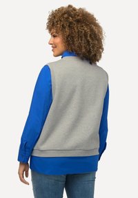 Grå ærmeløs sweater lagdelt over en lys blå langærmet skjorte, med en glat tekstur og ribbet kant, parret med denim jeans.