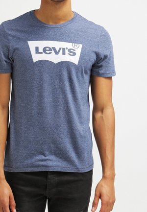 Man wearing a blue short-sleeve T-shirt with a white Levi's logo centered on the chest, paired with black pants.