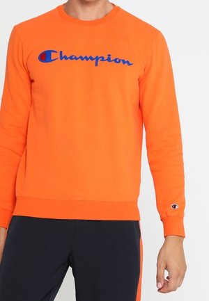 Oranje sweater met lange mouwen en een blauw "Champion"-logo op de borst, gedragen door een persoon in een zwarte broek met oranje strepen aan de zijkant.