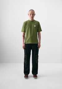 Oliven grøn kortærmet t-shirt med hvid stjerne logo, parret med sorte bukser med knælapper og brune sko med hvide såler.