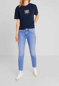 Námořnické bavlněné tričko s logem "TOMMY JEANS", kombinované se světle modrými džíny. Model má na sobě bílé vysoké tenisky. Neformální outfit.