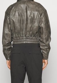 Person i en sliten brun skinnjacka med elastiskt midjeband och svarta byxor, sedd från ryggen mot en enfärgad bakgrund.