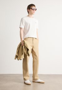 Tricou alb, pantaloni kaki și adidași albi cu accente bej. În mână, o jachetă de culoare maron deschis. Ochelari de soare cu ramă închisă la culoare.
