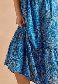 Modrá šaty s vzorom, ktoré majú voľný strih, zberný pás a jemnú textúru s plynulým lemom zvýrazneným zlatými bodkami.