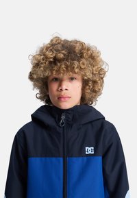 Niebieska i czarna kurtka z kapturem zapinana na zamek, z logo na piersi i gładką fakturą. Zaprojektowana dla młodzieży w prostym wzorze bloków kolorystycznych.