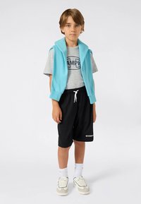 Világoskék ujjatlan kapucnis pulóver szürke "CHAMPION" logós póló fölött, fekete rövidnadrág, fehér zokni és szürke, díszítésekkel ellátott tornacipő.