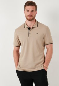 Beige rövidujjú póló, fekete gallérral és ujjkiemelésekkel, bal mellkason egy kis emblémával, puha szövettextúrával.