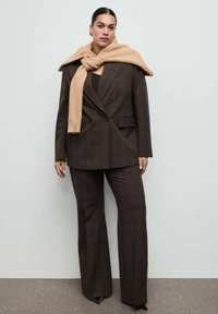 Kobieta w ciemnobrązowym, dwurzędowym garniturze z szerokimi spodniami oraz camelowym szalikiem przewiązanym na szyi, na jednolitym tle.