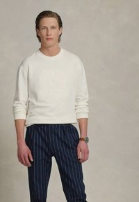 Sudadera albă cu design embossat, combinată cu pantaloni în dungi bleumarin. Include un ceas la încheietura stângă. Întreaga ținută este simplă și casual.