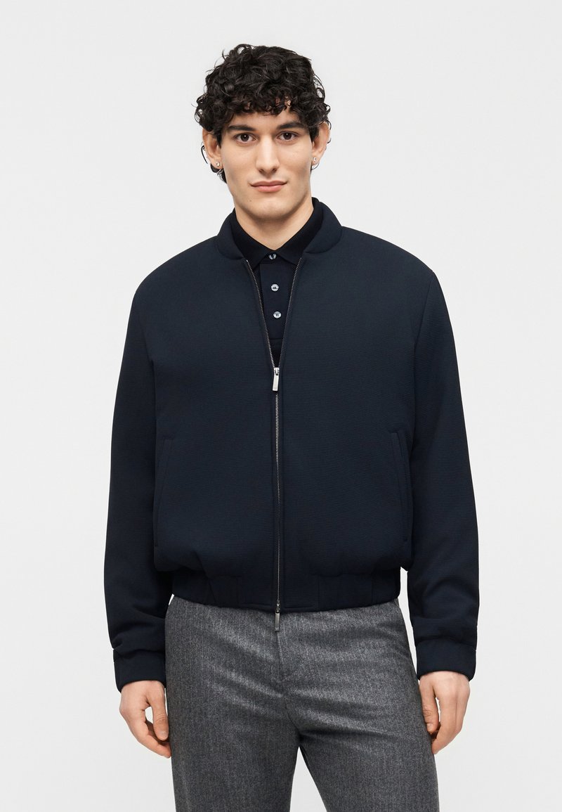 Νεαρός με σγουρά μαλλιά, ντυμένος με σκούρο ναυτ ικό μπουφάν bomber με φερμουάρ πάνω από μαύρο πουκάμισο με κουμπιά και γκρι ραμμένο παντελόνι.