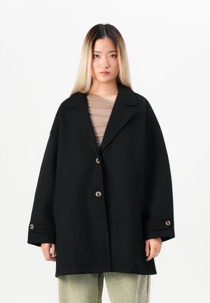 Jeune personne avec de longs cheveux dégradés, portant un manteau noir ample à boutons et un pantalon clair, se tenant devant un fond blanc.