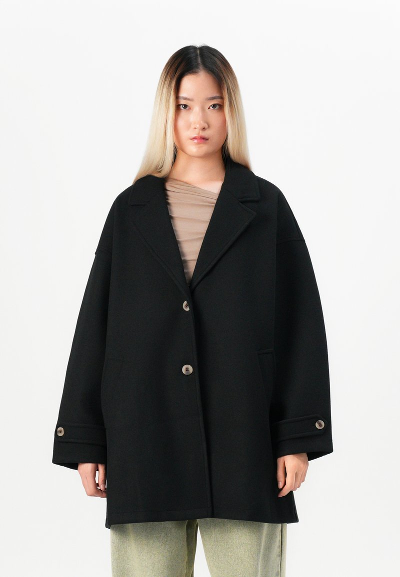 Young person with long ombre hair wearing an oversized black buttoned coat and light-colored pants, standing against a white background.
