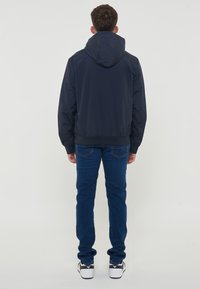 Marinblå hættejakke med elastiske manchetter og talje, parret med mørkeblå jeans og sorte sneakers. Stoffet fremstår let og glat.