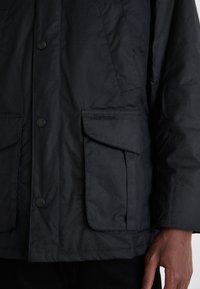Veste noire imperméable avec un bouton sur le devant, deux grandes poches poitrine et un logo 'Barbour' en relief sur la poche gauche.
