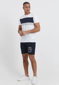 Fehér póló, navy és szürke vízszintes csíkozással, navy sorttal párosítva, amelyen egy fehér "Vintage" grafika található, fehér sportcipővel viselve.