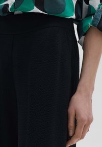 Pantalons noirs texturés à taille ajustée. Main visible reposant sur le côté, mise en valeur par une chemise à motifs colorés.