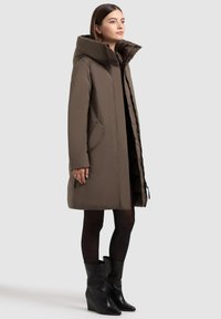 Bruine pufferjas met capuchon, ritsluiting, zijzakken en een rechte pasvorm. Gedragen over zwarte legging en sleehak enkellaarsjes.