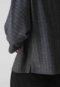 Blazer de lana gris con patrón de espiga que presenta un ligero vuelo en el dobladillo, mangas largas y una textura suave. Pantalones negros visibles.