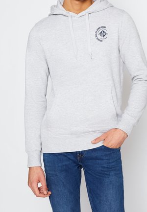 Mand iført en lysegrå hættetrøje med forlommer og blå jeans, hånd hvilende i lommen, logo trykt på venstre bryst.
