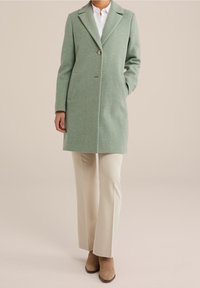 Vihreä villatakki, jossa lovikaulus, yksirivinen napitus ja kaksi sivutaskua. Yhdistetty kermaisen värisiin housuihin ja ruskeisiin nilkkureihin.